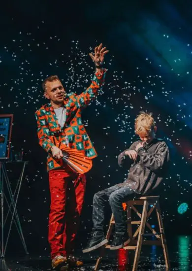 Kouzelník Mišuge v pestrém saku zvedá ruku v padajícím sněhu, vedle něj sedí malý divák. Vánoční speciál v Divadle Bolka Polívky. KULTINO*