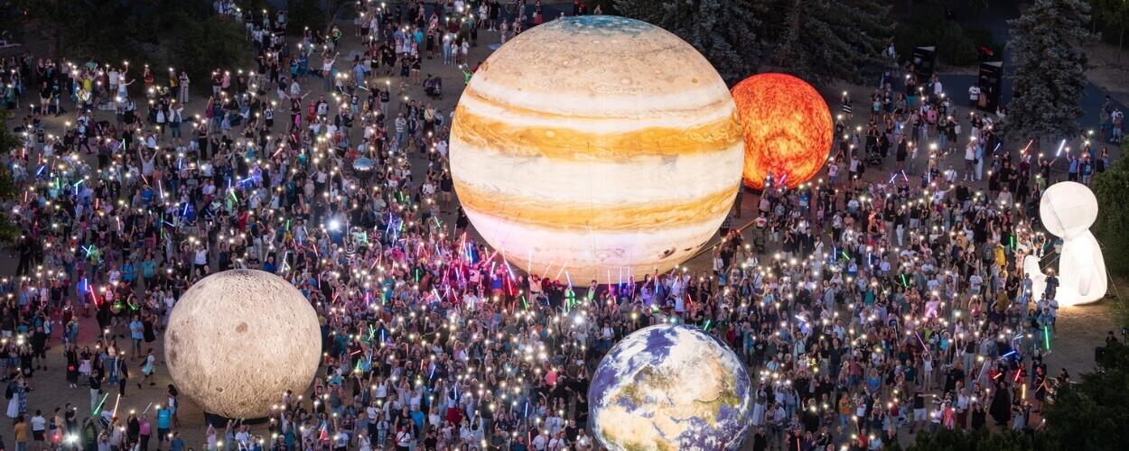 Obrovský dav návštěvníků se svítícími telefony obdivuje obří svítící modely planet, včetně Jupitera a Země, na brněnské Kraví hoře během Festivalu planet. KULTINO*