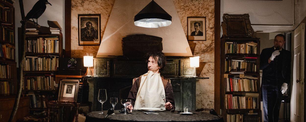 Představení Delikatesa od Mikro teatro. Zaskočený herec s ubrouskem kolem krku sedí u prostřeného stolu v netradičním divadelním prostoru opuštěného brněnského bytu plného knih. KULTINO*