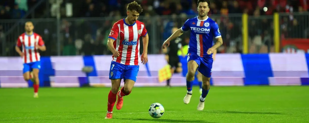 Dramatický souboj o míč během vyostřeného brněnského fotbalového derby. Hráči favorizované Zbrojovky a posíleného Atrisu bojují na trávníku plném emocí před burácejícími tribunami. KULTINO*
