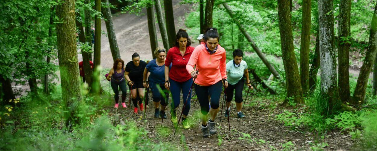 Skupina lidí stoupající lesním terénem s chodeckými holemi při lekci Nordic walkingu. KULTINO*