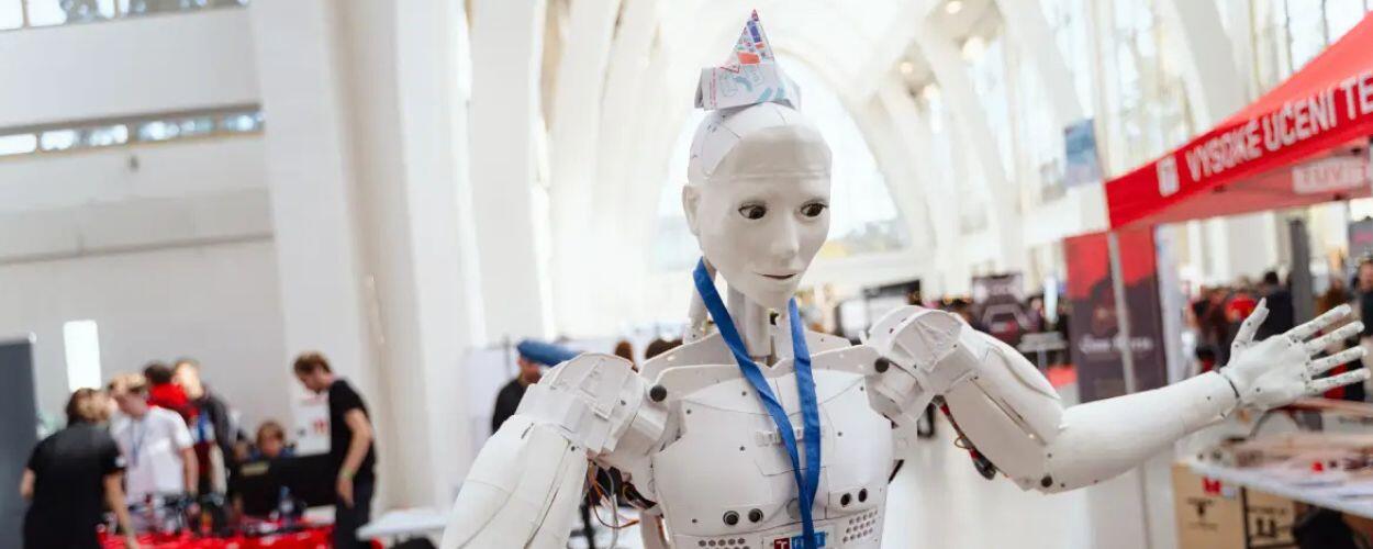 Bílý humanoidní robot s papírovou čepicí a modrou šňůrkou na krku mává mezi stánky makerů na festivalu Maker Faire Brno. KULTINO*