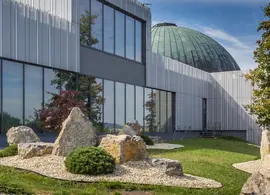 Moderní budova Hvězdárny a planetária Brno s prosklenou fasádou a zelenou kopulí, před ní skalka v parku na Kraví hoře. KULTINO*