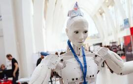 Bílý humanoidní robot s papírovou čepicí a modrou šňůrkou na krku mává mezi stánky makerů na festivalu Maker Faire Brno. KULTINO*